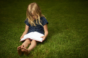 Fototapeta premium Cute Little Blond Girl Reading Book Outside on Grass