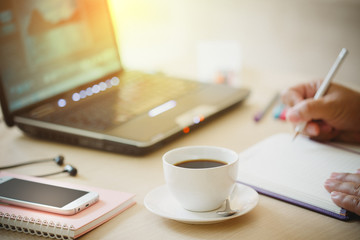 close up cup of coffee and smart phone  with hand of business man using laptop computer and write notebook on wooden desk office with morning light. vintage filter effect.