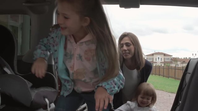 Mother and two her little daughters going to the car: woman lifting up laughing girl and putting her into baby seat