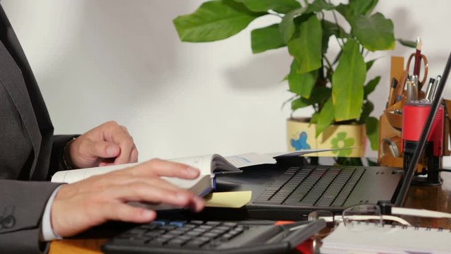 Man in formal suit sitting at the table makes calculations, take pen and makes notes in notebook. Organized working place in office, plant, laptop, glasses on table. Closeup. 4K Ultra HD.
