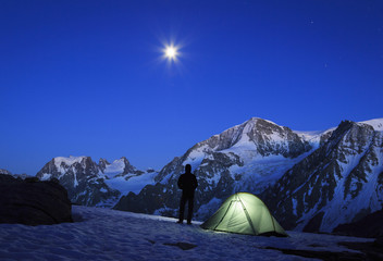 Mountaineer beside his tent, looking at the moon rising above the mountains in Wallis, Switserland.