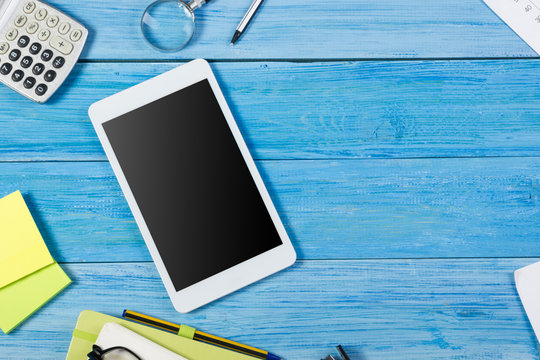 Tablet pc, empty blank screen on office table desk with set of colorful supplies, note pad, cup, pen, flower blue background. Top view and copy space for ad text