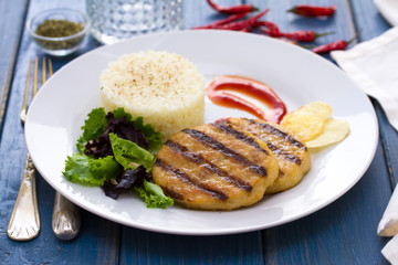grilled hamburger with rice, salad and chips on plate on blue background