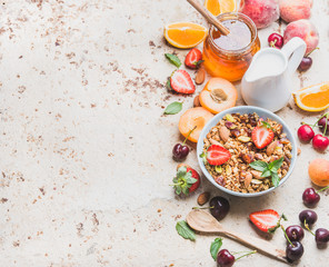 Healthy breakfast ingredients. Oat granola in bowl with nuts, strawberry and mint leaves, milk in pitcher, honey in glass jar, fresh fruits and berries on light concrete background, top view
