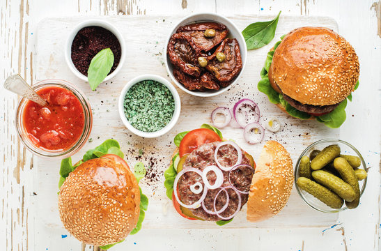 Homemade beef burgers with onion, pickles, fresh vegetables, sun-dried tomatoes and tomato sauce on serving wooden board over white painted background. Top view, horizontal composition