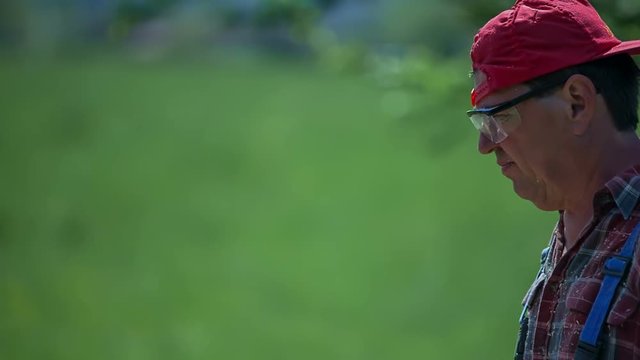 A joiner looks very dedicated and focused when he is cutting wood in the garden. It is a nice summer day. He is wearing his hat backwards.
