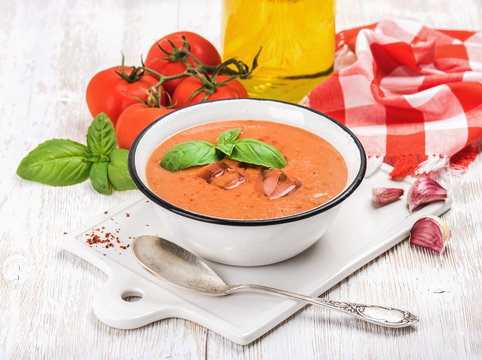 Cold gazpacho soup in bowl with ice, hot pepper and basil served with fresh tomotoes on ceramic board over white wooden background, selective focus, horizontal composition