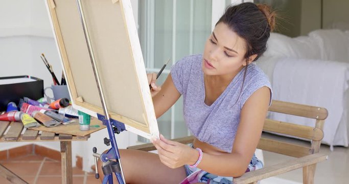 Attractive female artist working on a canvas concentrating as she paints in a detail on an outdoor patio at home