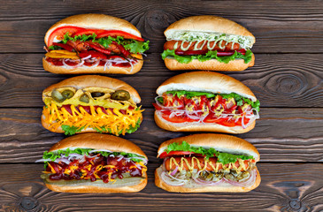 A lot of big delicious hot dogs with sauce and vegetables on wooden background. Their assorted hot...