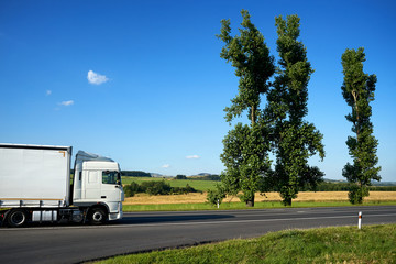 Fototapeta na wymiar White truck driving on asphalt road past the two tall poplars in the countryside under a blue sky.