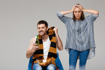 Man drinking beer, watching football, upset woman standing behind.