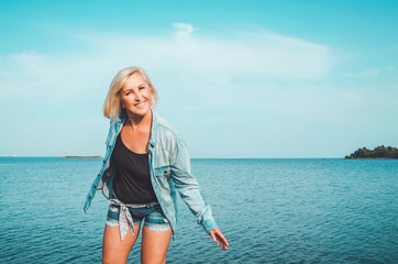 Tanned healthy middle aged woman with denim clothes having fun. Active, enjoy concept on sunny summer day, outdoors. Happiness carefree female posing in dance on beach, blue sky background with sea.