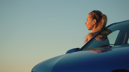 Beautiful young girl standing at the car into the sunset