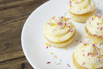 A plate of birthday cupcakes with vanilla frosting and sprinkles on a wooden background