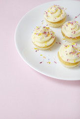 A plate of birthday cupcakes with vanilla frosting and sprinkles on a pink background