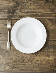 An empty white dinner plate and fork on a rustic wooden background