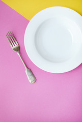 An empty white dinner plate and fork on a two toned pink and yellow background
