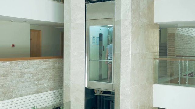 A female is standing in glass transparent lift that is going up and down. Elevator makes the commute to higher floor very easy. Lifts are best for a disabled person.