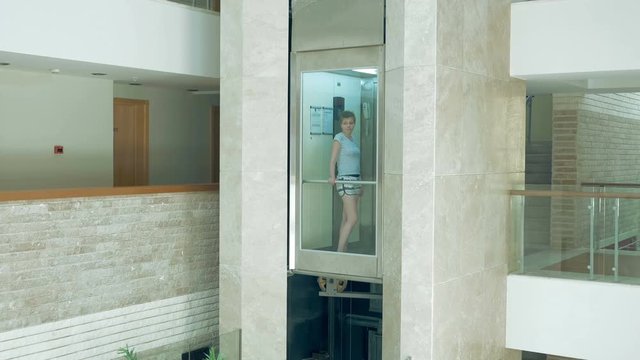 A female is standing in glass transparent lift that is going up and down. Elevator makes the commute to higher floor very easy. Lifts are best for a disabled person.