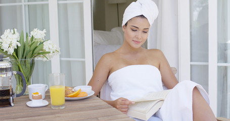 Cheerful young adult woman wrapped in white bath towel reading book at table with flowers  coffee  juice and fruit