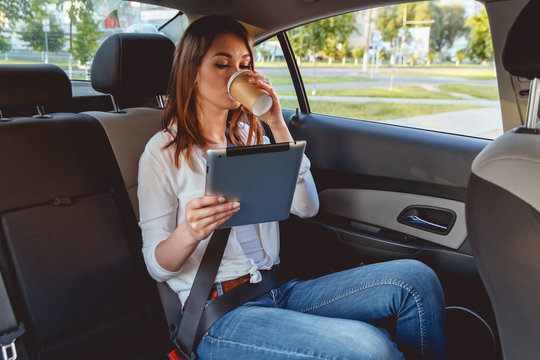 Young, Beautiful Woman Sitting In The Back Seat Of The Car With A Tablet In Hand And Drinking Coffee