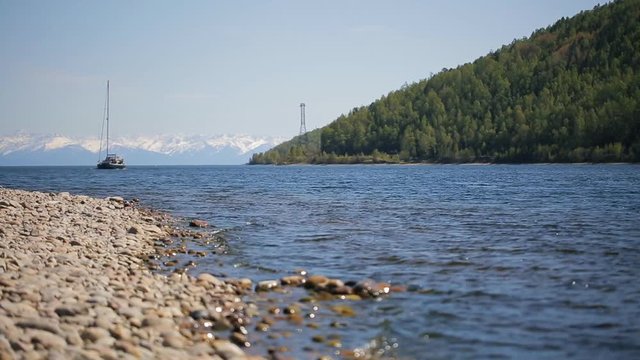 A sailing boat moves away from pebbly shore in background of a chain of mountains and steep forest.