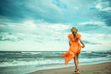 A young slender woman in orange dress is walking barefoot towards the storming sea. The train of her dress is waving. Cool wind in her hair. Outdoor shot. Copy-space.