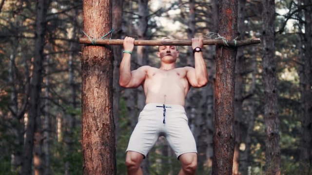 Handsome and strong bodybuilder doing pull-ups on handmade horizontal bar in forest