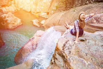 Happy woman sitting on the rock edge near waterfall enjoying stunning view, filtered with sun flares