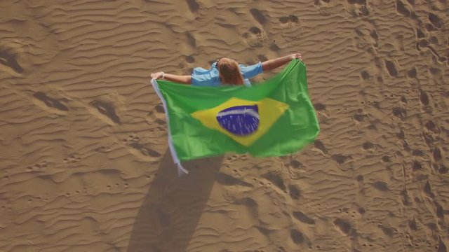 Aerial shot of a woman on the sand holding flag of Brazil overhead. National symbol fluttering in strong wind. Rio Olympic Games 2016