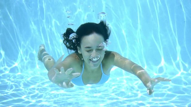 Happy young woman smiling underwater as she enjoys a swim in a sparkling blue swimming pool facing the camera with copy space