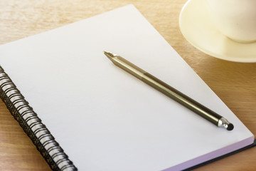 Blank notebook on the wooden desk background with a pen on top and a coffee cup the right side.