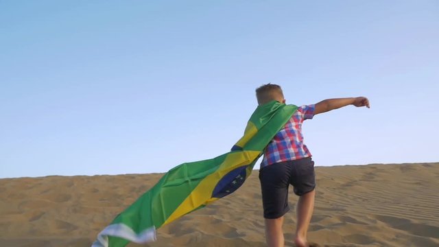 Slow motion shot of a boy with flag of Brazil tied as cape running away on the sand and celebrating the victory standing on sky background. Summer Olympic Games in Rio 2016
