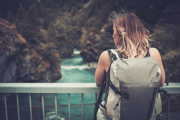 Fototapeta premium Woman hiker with backpack standing on the bridge over a wild mountain river.