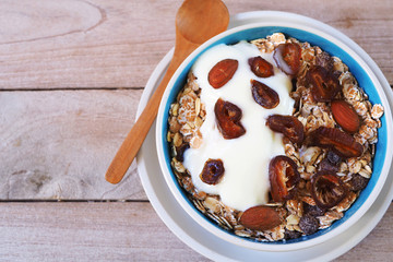 Top view of a healthy bowl of Muesli with whole grain, oats, dried fruit, nuts and date fruit with yogurt on a wooden table.