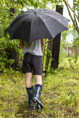 Child in rubber boots goes on a footpath to the wicket through a puddle in the rain under a black...