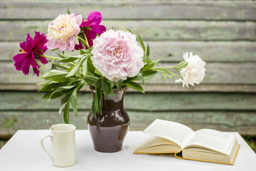 Vase the beautiful peonies, a cup with hot drink, the open book on a table covered with a tablecloth, outdoor, against a background the old wooden wall with peeling paint lovely summer afternoon