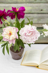 Vase of beautiful peony and the open book on a table covered with a tablecloth, outdoor, against a background the old wooden wall with peeling paint  lovely summer afternoon