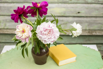 Vase of beautiful peony and a closed book on a table covered with a tablecloth, outdoors, against a background the old wooden wall with peeling paint  lovely summer afternoon