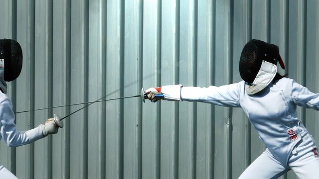 Slow motion fencing duel clip with two fencers fighting in front of a corrugated metal wall.
