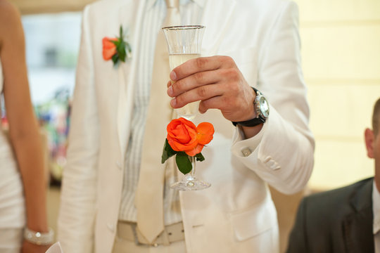 Groom in white suit holds a champagne fluet decorated with orang