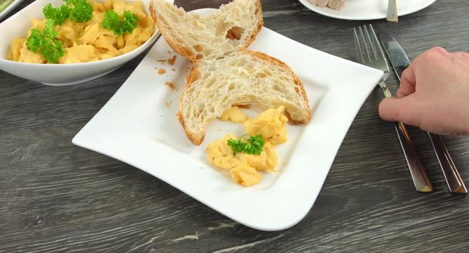 4K Clip eines Fr&uuml;hst&uuml;ckes. Ein aufgeschnittenes H&ouml;rnchen und etwas R&uuml;hrei liegen auf einem eckigen weissen Teller. Eine Frau belegt das H&ouml;rnchen mit frischem R&uuml;hrei.