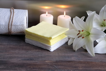 White and yellow paper napkins,paper towel on wooden table against steel background with lily flowers and burning candles. Concept of spa and body care