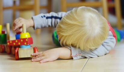 Little blond boy lying on the floor is passionated about  playing with wooden locomotive