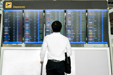 Asian young business man with luggage waiting for travel in airport near flight timetable.