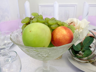 Fruit in a glass vase on a festive table. Apples and grapes on the dining table as a dessert during the meal. Healthy vegetarian food.