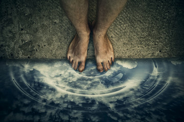 Surrealistic image with male bare legs standing on the edge, in front of water pothole reflecting a cloudy sky.