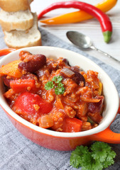 chili con carne mit chillies, frischem Gemüse, Petersilie und Gewürzen in einer kleinen orangenen Schüssel mit Löffel und Brot auf einem grauen Tischtuch