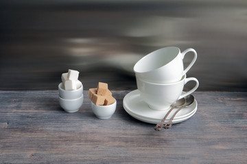 White china porcelain dishes on a wooden table with flower of lily. Two cups stacked with tea spoons. Special light. Toned image.