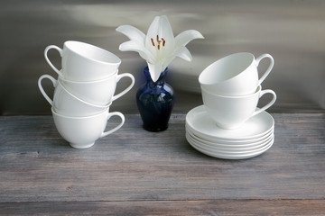 White china porcelain dishes on a wooden table with flower of lily. Six cups stacked. Special light. Toned image.